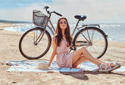 Luchtige Zomerjurken: vrouw met roze jurk aan op strand, met achter haar haar fiets