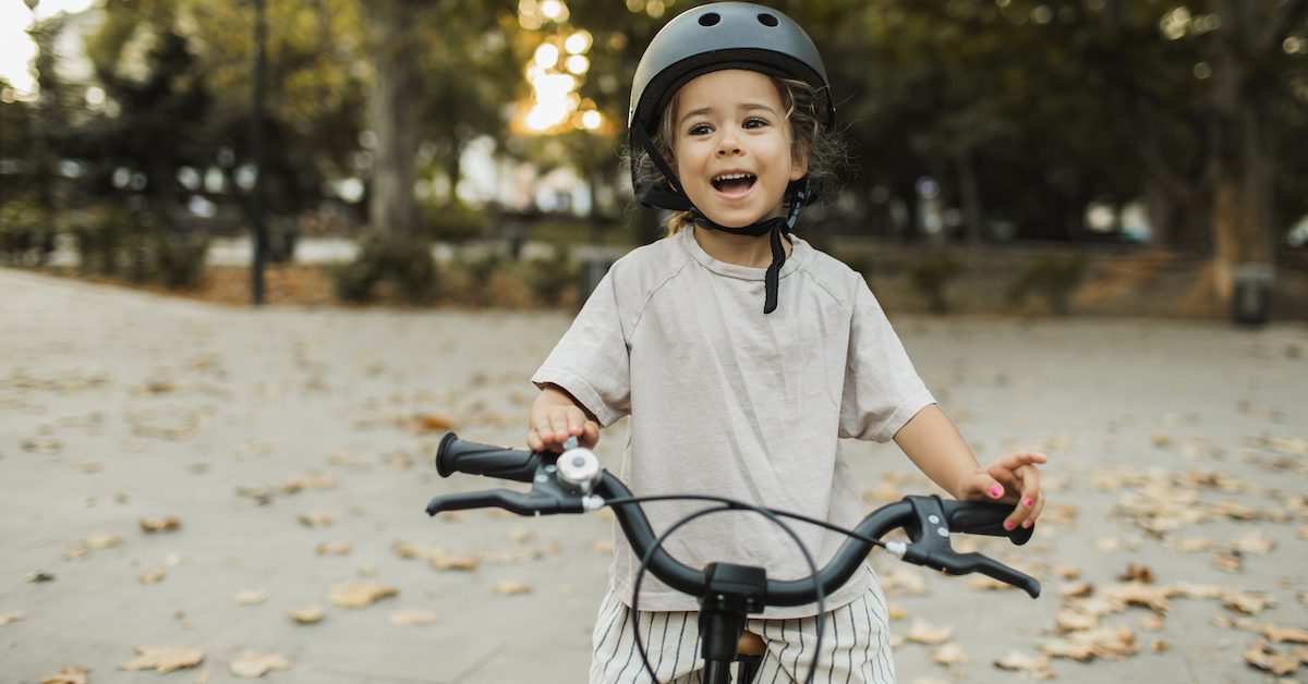 Cheerful Child Riding A Bike In Helmet In City Park. Happy Childhood