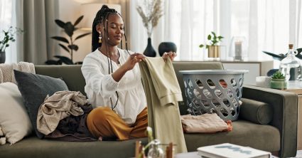 Laundry, Housework And A Black Woman Folding Washing On A Sofa In The Living Room Of Her Home To Tidy. Smile, Relax And A Happy Young Housewife Cleaning Her Apartment For Housekeeping Or Hygiene