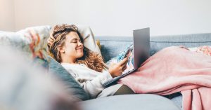 Woman Wrapped In Blanket Using Laptop On Sofa At Home.