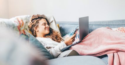 Woman Wrapped In Blanket Using Laptop On Sofa At Home.