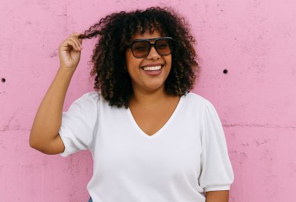 Plussize Kledingtrends: vrouw met wit shirt en zonnebril op, roze muur als achtergrond