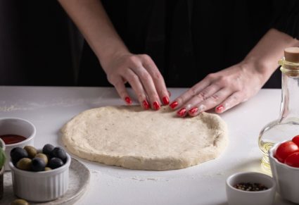 Vrouw die deeg uitrolt voor zelf pizza bakken, omringd door olijfolie, tomaten en olijven op het aanrecht.